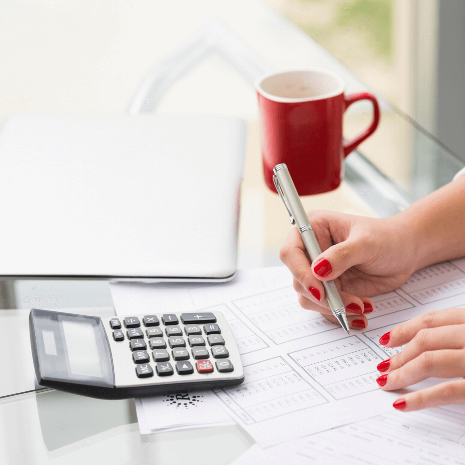 A desk with a report, calculator, closed laptop, and coffee mug. You see a womans hands holding a pen.